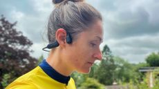Hannah is wearing the Haylou PurFree headphones over her right ear. She has her hair tied up and has a yellow jersey on. There is greenery and a grey sky in the background
